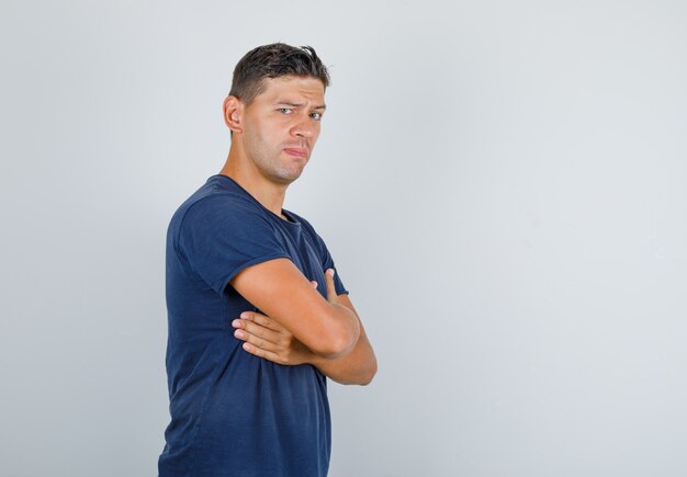 Hombre joven cruzando las manos sobre el pecho con cejas fruncidas en camiseta azul oscuro y mirando confiado.