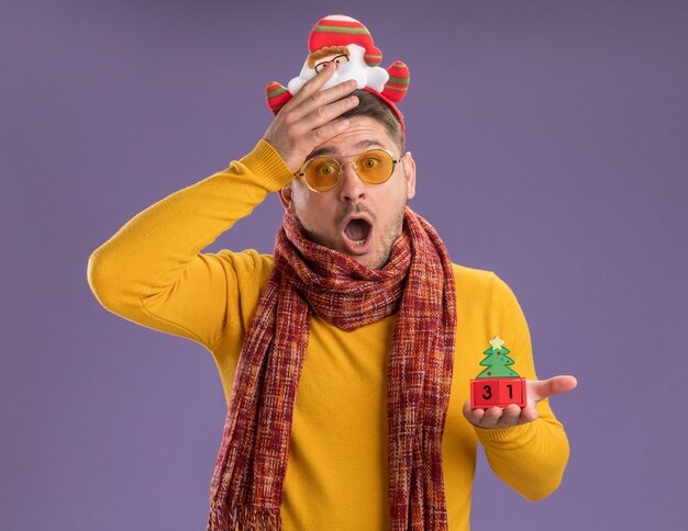 Hombre joven de cuello alto amarillo con bufanda cálida y gafas con borde divertido con santa en la cabeza mostrando cubos de juguete con fecha de año nuevo asombrado y sorprendido de pie sobre la pared púrpura
