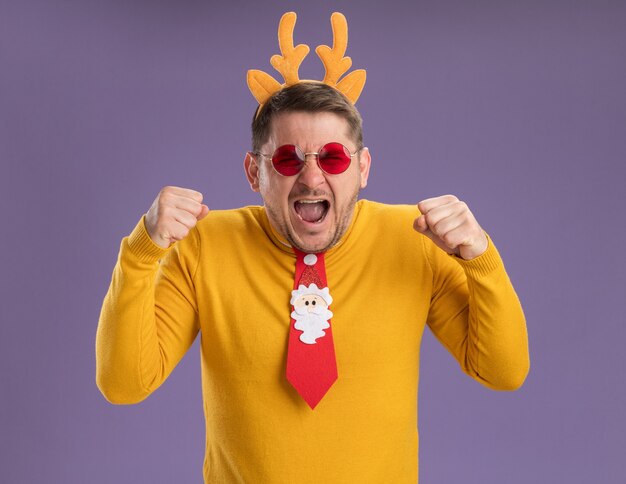 Hombre joven con cuello alto amarillo y gafas rojas con corbata roja divertida y borde con cuernos de venado en la cabeza gritando con expresión agresiva apretando los puños sobre fondo púrpura