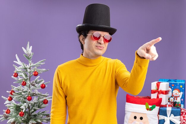 Hombre joven con cuello alto amarillo y gafas con sombrero negro mirando a un lado disgustado apuntando con el dedo índice a algo de pie junto a un árbol de Navidad y presenta sobre fondo púrpura