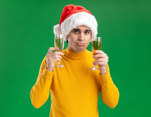Hombre joven de cuello alto amarillo y gorro de Papá Noel sosteniendo dos copas de champán mirando a la cámara con expresión triste de pie sobre fondo verde