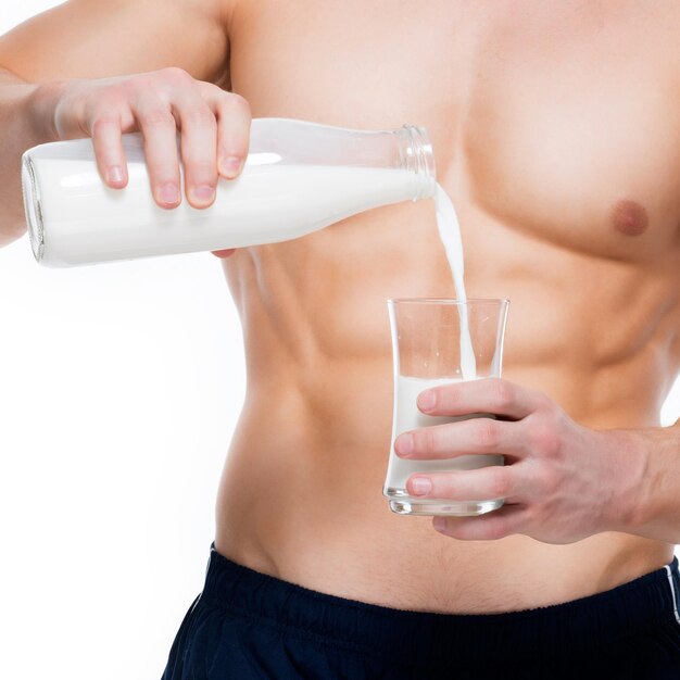 Hombre joven con cuerpo perfecto vertiendo leche en un vaso - aislado en la pared blanca.