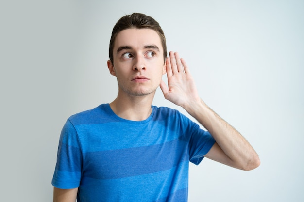 Hombre joven curioso que lleva a cabo la mano cerca de la oreja