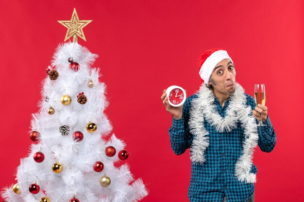 Hombre joven curioso con sombrero de santa claus y sosteniendo una copa de vino y reloj de pie cerca del árbol de Navidad