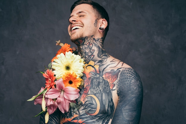 Hombre joven descamisado alegre con orejas perforadas con decoración de flores en su cuerpo tatuado