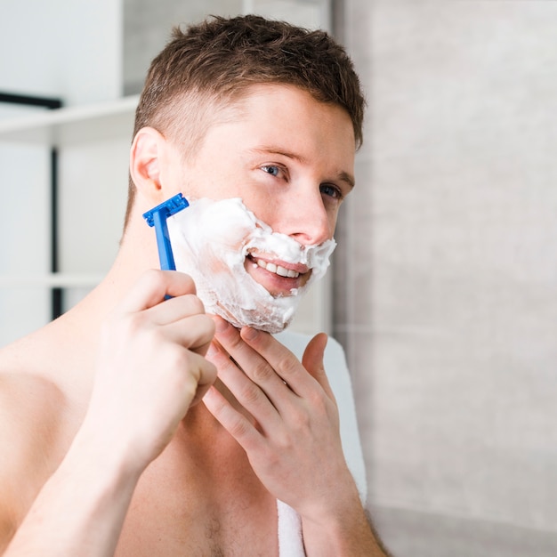 Hombre joven descamisado sonriente que afeita con la maquinilla de afeitar azul
