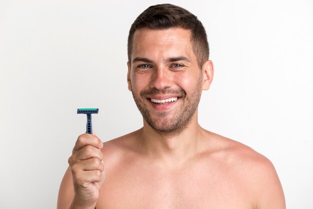 Hombre joven descamisado sonriente que sostiene la maquinilla de afeitar que se opone al fondo blanco