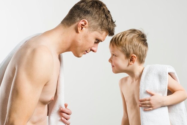 Hombre joven descamisado y su hijo con la toalla doblada en su hombro mirándose el uno al otro contra el fondo gris
