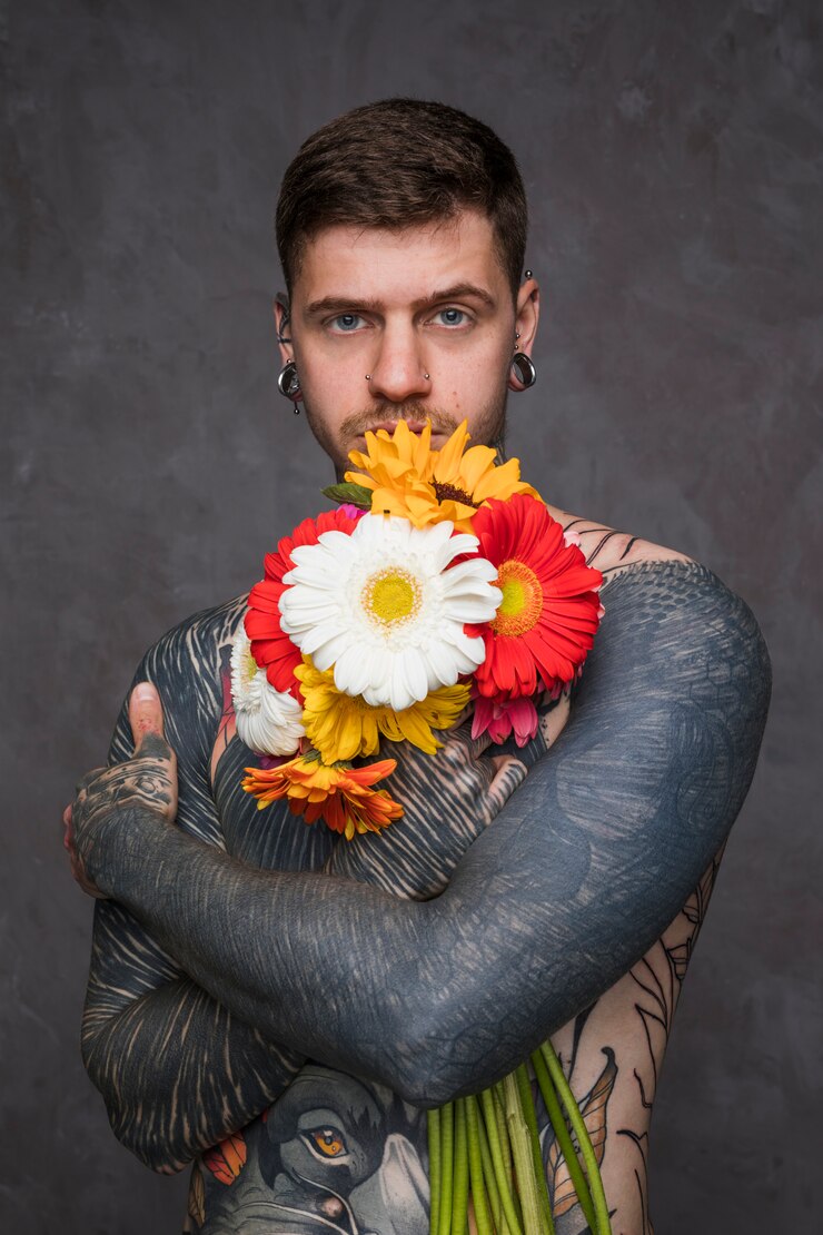 Foto gratuito hombre joven descamisado con tatuado en su cuerpo que sostiene las flores hermosas del gerbera en la mano que se opone al fondo gris