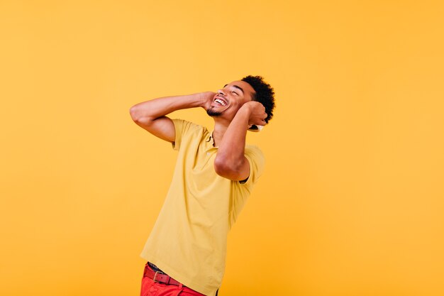 Hombre joven dichoso en camiseta brillante de moda riendo. Foto interior de chico africano emocional sonriendo con los ojos cerrados.