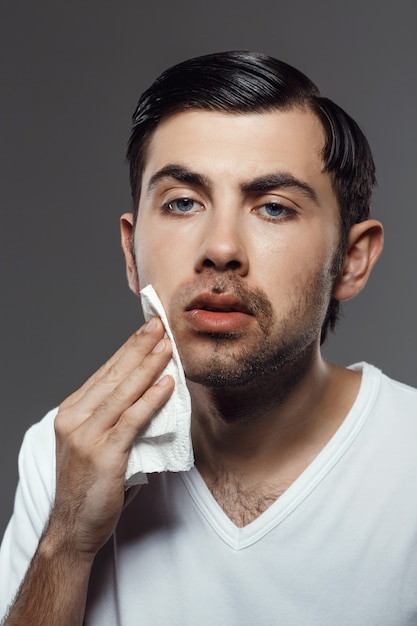 Hombre joven disgustado tocando la cara después de afeitarse sobre la pared gris