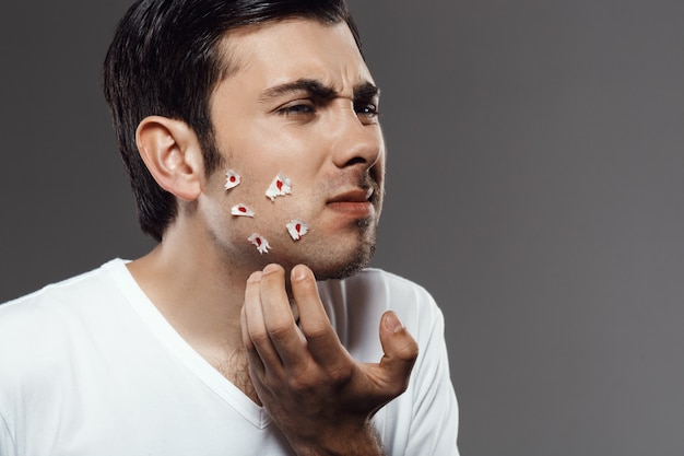 Hombre joven disgustado tocando la cara después de afeitarse sobre la pared gris