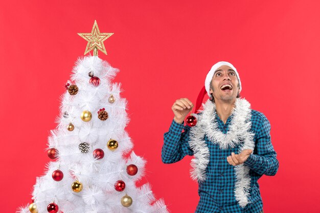 Hombre joven divertido con sombrero de santa claus en una camisa azul a rayas y sosteniendo accesorio de decoración