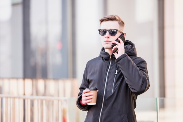 Hombre joven elegante con las gafas de sol usando el teléfono móvil que sostiene la taza de café para llevar