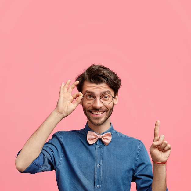 Foto gratuita hombre joven elegante positivo con peinado de moda y barba de varios días, vestido con camisa de mezclilla con pajarita, tiene una expresión alegre, apunta hacia arriba contra la pared rosa, mantiene la mano en el borde de las gafas