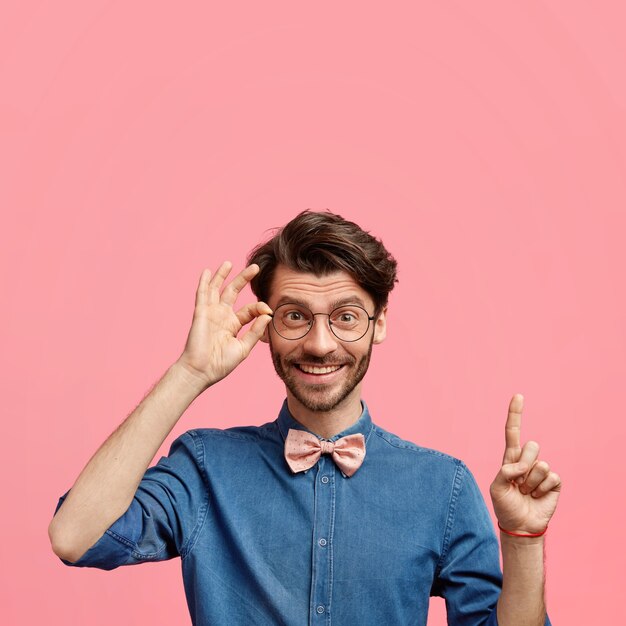 Hombre joven elegante positivo con peinado de moda y barba de varios días, vestido con camisa de mezclilla con pajarita, tiene una expresión alegre, apunta hacia arriba contra la pared rosa, mantiene la mano en el borde de las gafas