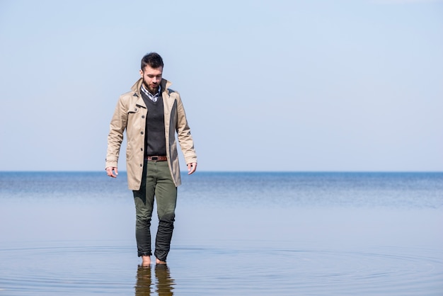 Hombre joven elegante que camina en el agua de mar poco profunda contra el cielo azul