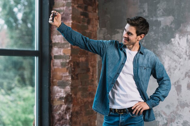 Hombre joven elegante que presenta delante de la ventana que toma el selfie del teléfono móvil