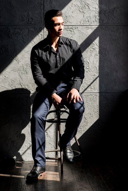 Hombre joven elegante que se sienta en taburete en luz del sol contra la pared gris