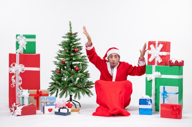 Hombre joven emocionado confundido vestido como Papá Noel con regalos y árbol de Navidad decorado sentado en el suelo sobre fondo blanco.