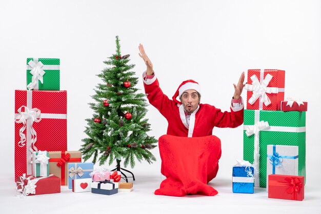 Hombre joven emocionado confundido vestido como Papá Noel con regalos y árbol de Navidad decorado sentado en el suelo sobre fondo blanco.