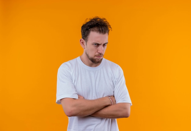Hombre joven enojado con camiseta blanca cruzando las manos en la pared naranja aislada