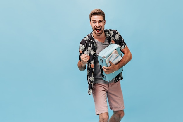 Hombre joven con estilo alegre con peinado fresco y barba en camisa de verano moderna y pantalones cortos beige con maleta azul y se regocija