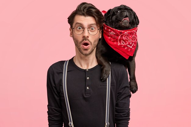Hombre joven estupefacto lleva su perro de raza negra en el cuello, vestido con una camisa de moda con tirantes, nota algo sorprendente, aislado sobre una pared rosa. Concepto de buenas relaciones.
