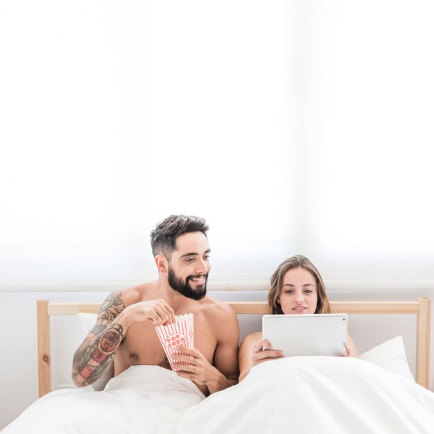 Hombre joven feliz con las palomitas que miran a su novia que usa la tableta digital en cama