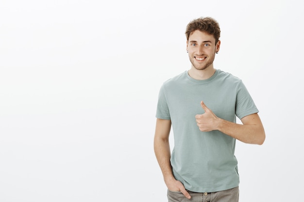 Hombre joven feliz positivo en aretes, sonriendo ampliamente sosteniendo la mano en el bolsillo y mostrando los pulgares para arriba