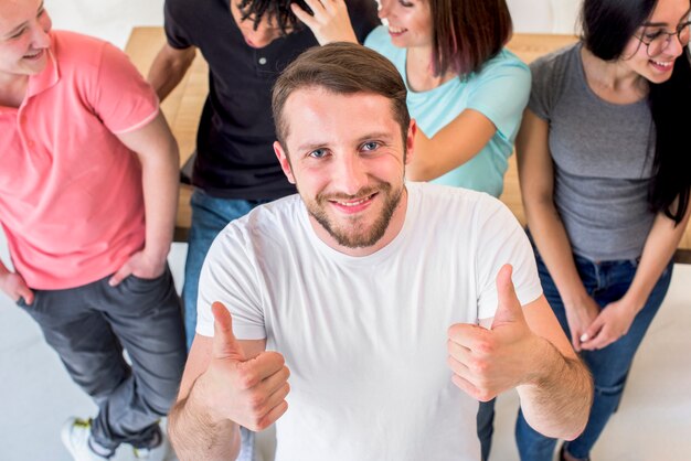 Hombre joven feliz que se coloca con los amigos que muestran el gesto del thumbup que mira la cámara