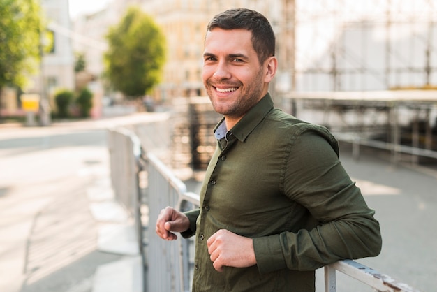 Foto gratuita hombre joven feliz que se coloca cerca de la verja metálica que mira la cámara