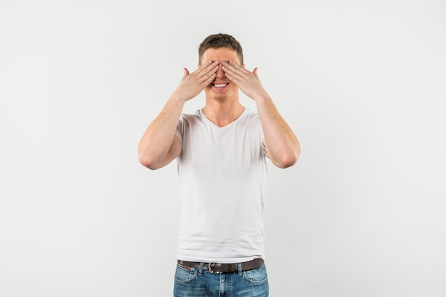 Hombre joven feliz que cubre sus ojos con dos manos contra el fondo blanco