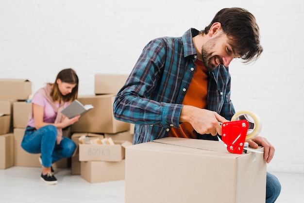 Hombre joven feliz que embala la caja de cartón y su esposa en el fondo