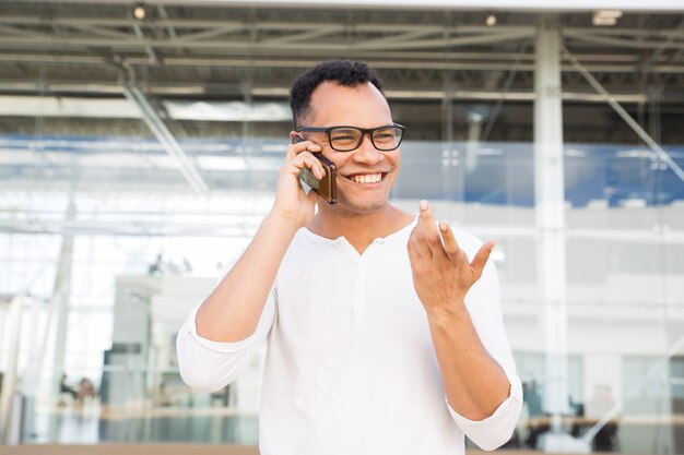 Hombre joven feliz que habla en smartphone y que gesticula al aire libre