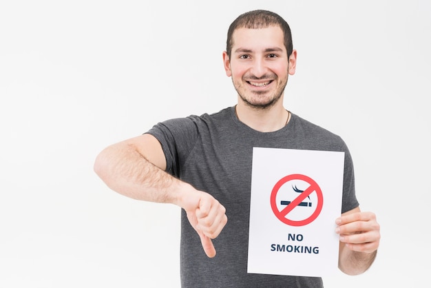 Hombre joven feliz que lleva a cabo la muestra de no fumadores que muestra el pulgar abajo aislado en el contexto blanco