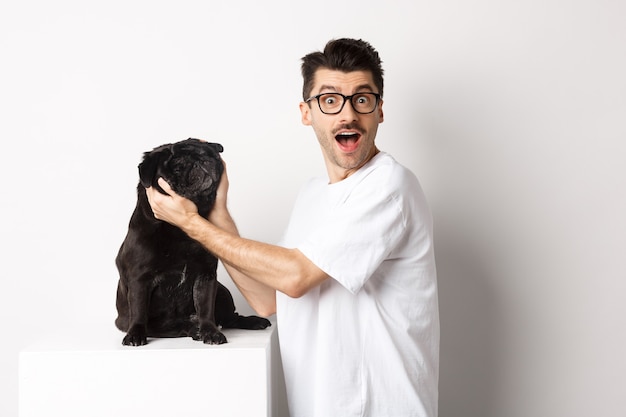 Hombre joven feliz que le muestra la cara linda de su pug. Dueño de perro amando a su mascota, de pie sobre fondo blanco.