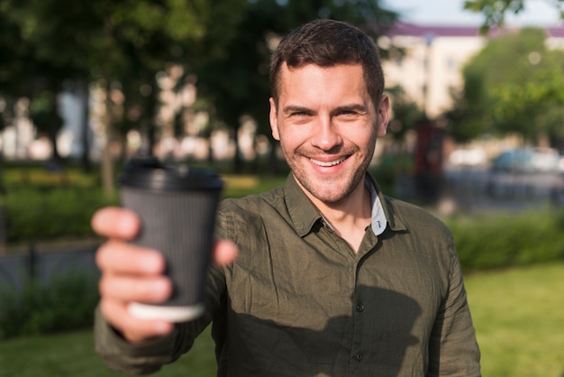 Hombre joven feliz que muestra la taza de café disponible en el parque