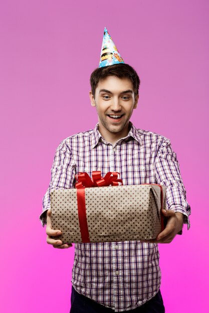 Hombre joven feliz que sostiene el regalo de cumpleaños en caja sobre la pared púrpura.