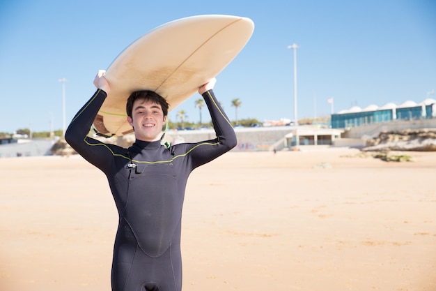 Hombre joven feliz que sostiene la tabla hawaiana en la cabeza en la playa soleada