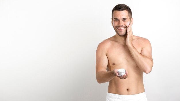 Hombre joven feliz que usa la crema en su cara contra la pared blanca