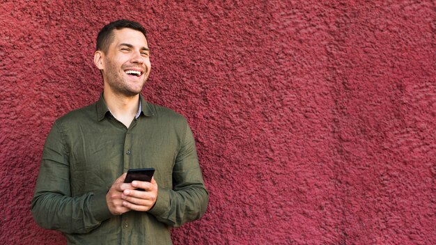 Hombre joven feliz del rastrojo que sostiene el teléfono móvil que se coloca cerca de la pared áspera