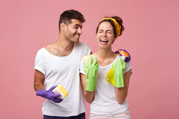 Hombre joven feliz reconfortante mujer estresada desesperada en guantes protectores que no tiene ganas de lavar los platos. Apuesto hombre positivo riéndose de su novia llorando triste que odia limpiar