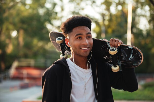 Hombre joven feliz del skater que sostiene el monopatín