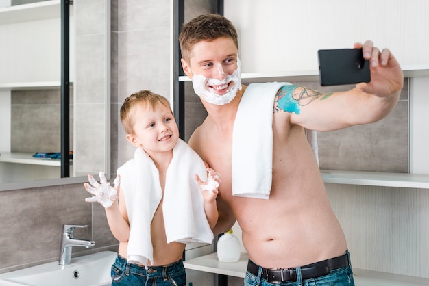Hombre joven feliz con su hijo en el baño tomando selfie en teléfono móvil