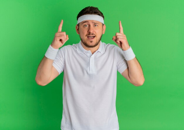 Hombre joven fitness en camisa blanca con diadema apuntando hacia arriba con los dedos índices mirando hacia el frente sorprendido de pie sobre la pared verde