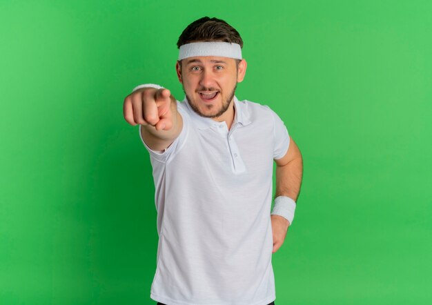 Hombre joven fitness en camisa blanca con diadema apuntando con el dedo hacia el frente sonriendo alegremente de pie sobre la pared verde