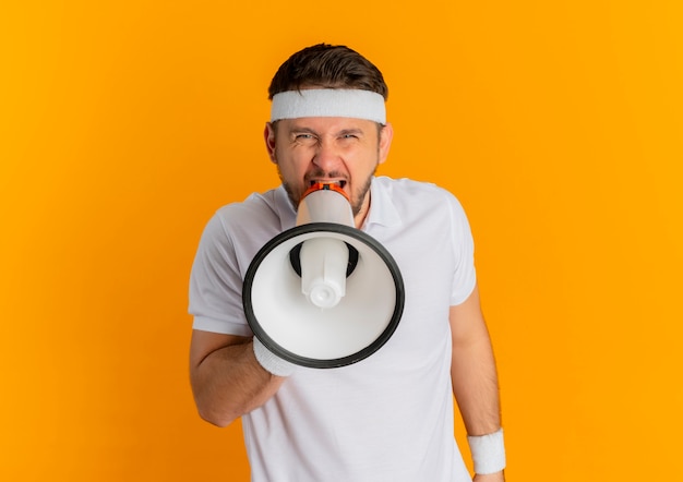 Hombre joven fitness en camisa blanca con diadema gritando al megáfono con expresión agresiva de pie sobre la pared naranja