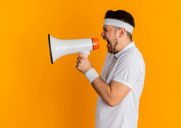 Hombre joven fitness en camisa blanca con diadema gritando al megáfono con expresión agresiva de pie sobre la pared naranja