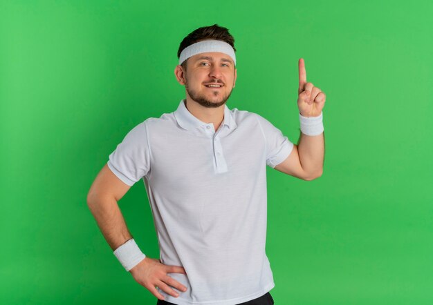 Hombre joven fitness en camisa blanca con diadema mirando al frente sonriendo confiado apuntando con el dedo hacia arriba de pie sobre la pared verde